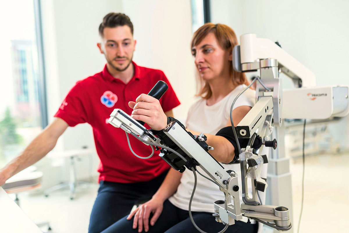 A woman practises arm movements on a rehabilitation device under a therapist’s supervision.